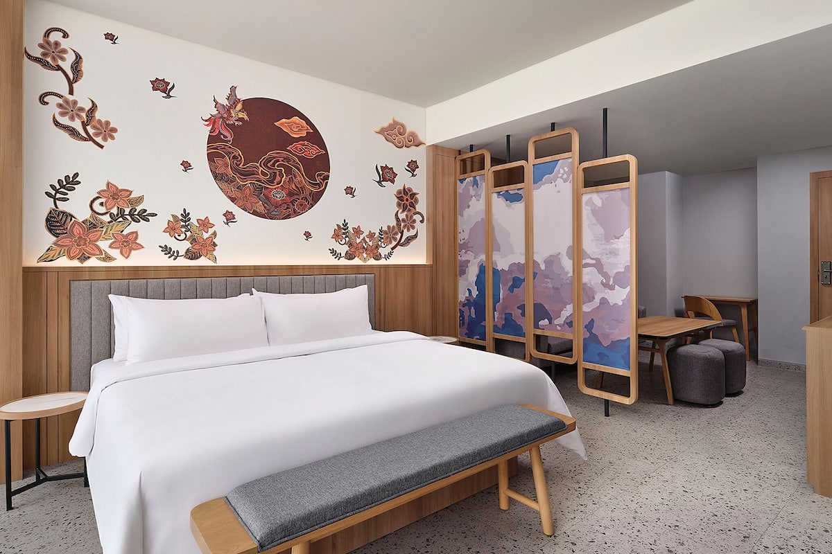A hotel room with a bed, a bench, a desk, and a partition with artwork.