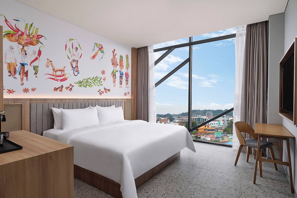 A hotel room with a large bed, a desk, a chair, and a city view through large windows.