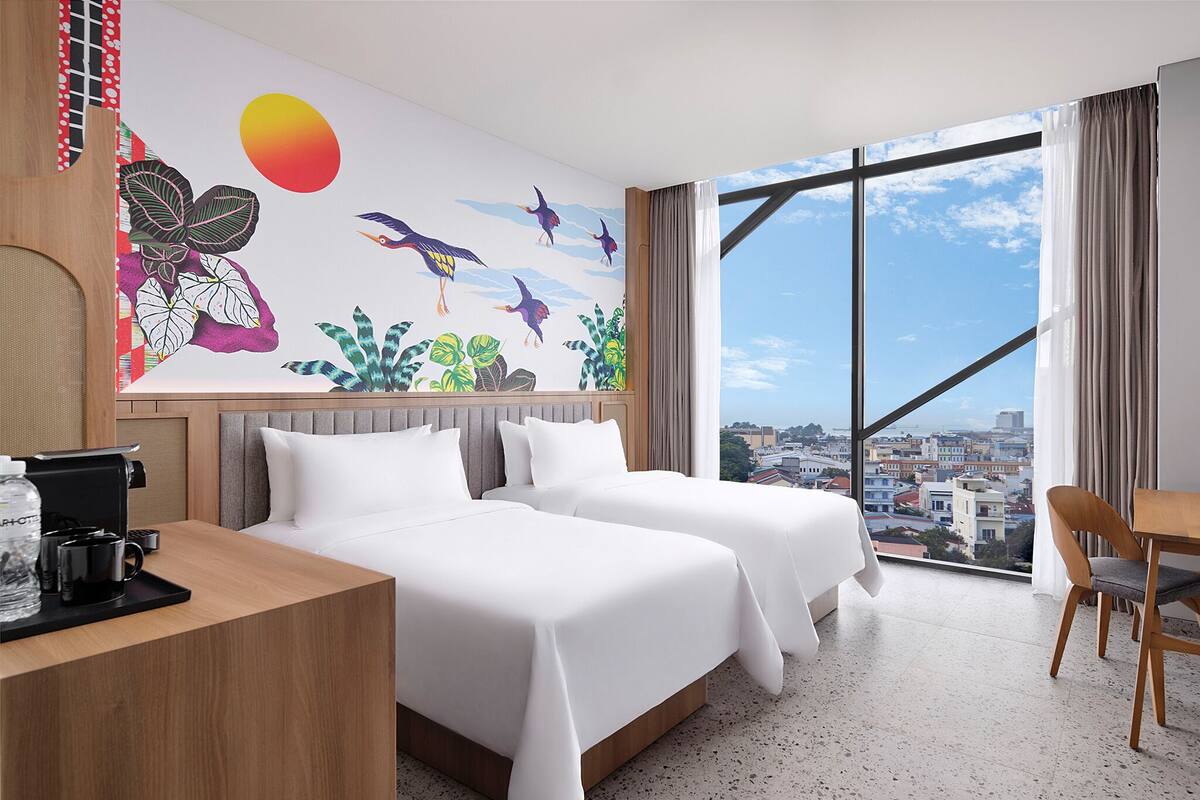 A hotel room with two beds, a desk with a coffee maker, a view of the city through large windows, and a mural with birds and plants.