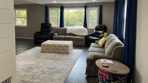 Amplia sala de estar con un sofá modular grande, una chaise lounge, un pequeño otoman y una mesa de centro redonda. Hay dos ventanas con cortinas azules, un techo con iluminación empotrada y vistas a árboles fuera.