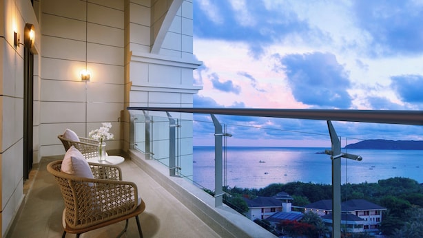 Un balcon agrémenté de fauteuils en osier et d’une petite table ornée de fleurs, d’où l’on peut admirer un plan d’eau et un paysage lointain.