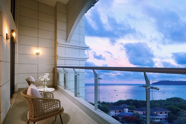 A balcony with wicker chairs and a small table with flowers, overlooking a body of water and distant land.