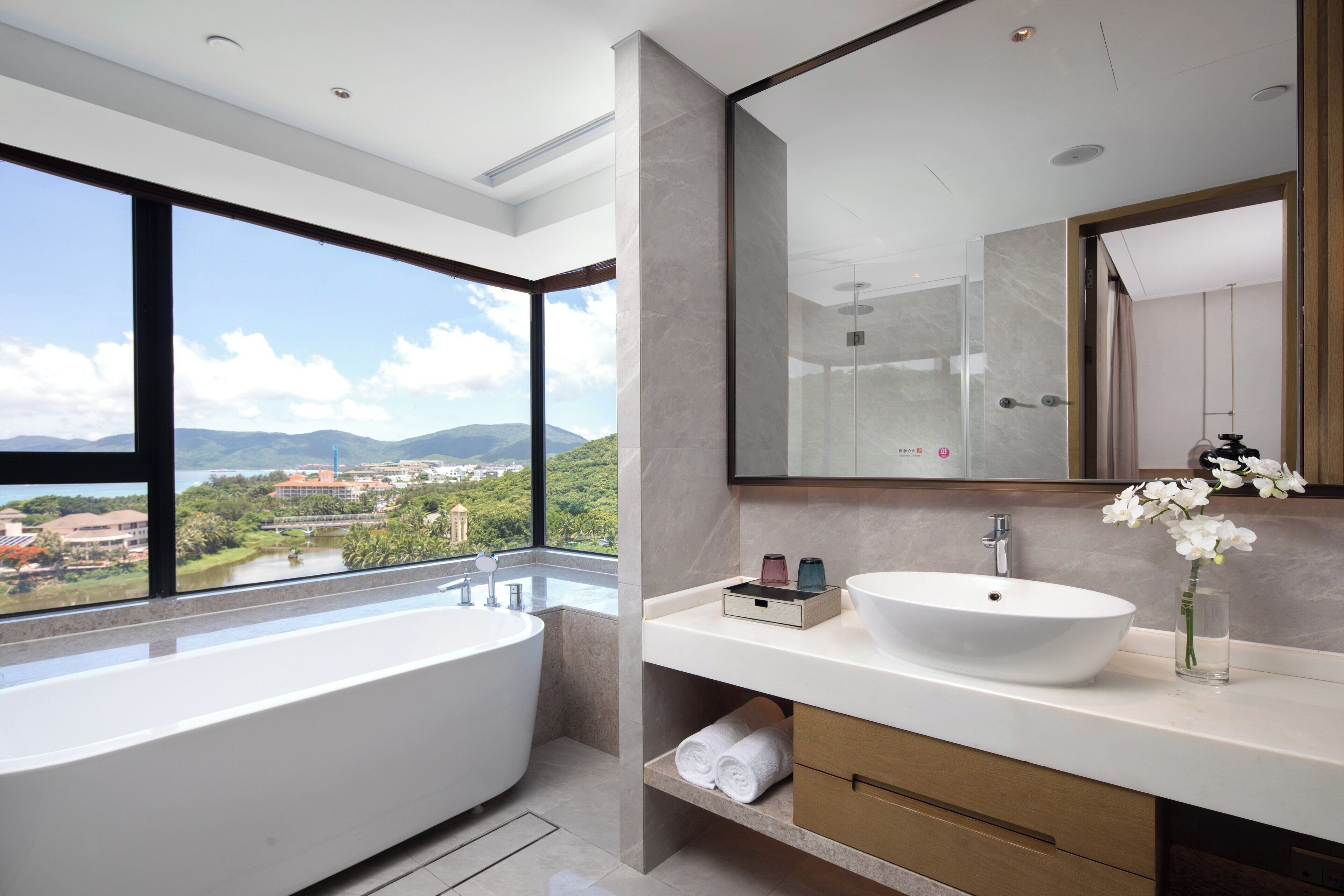 A modern bathroom with a large bathtub, a white sink, and a large mirror. There is a view of a city and mountains through a large window.