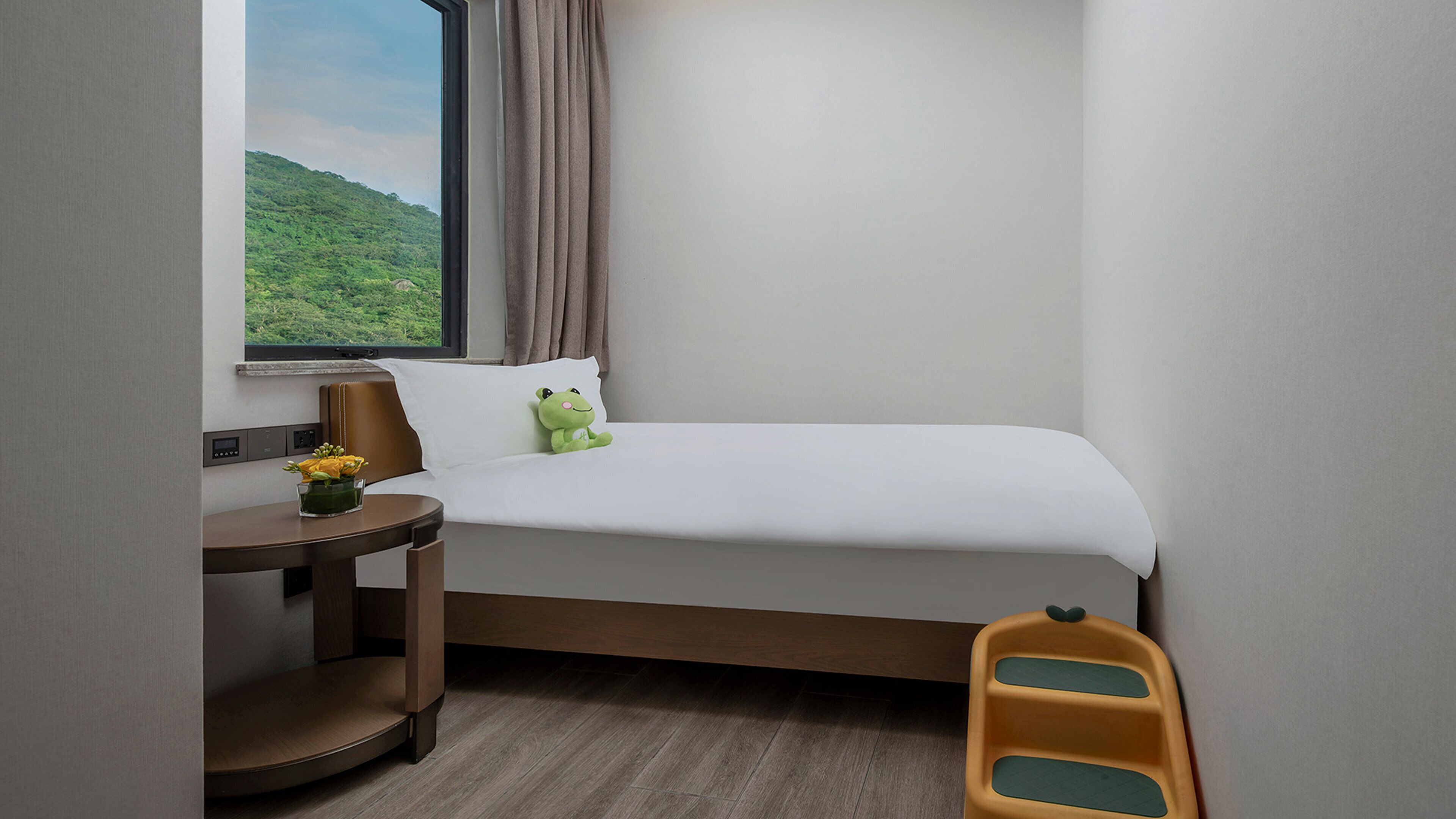 A hotel room with a bed, a small table with a vase of flowers, a window with a view of greenery, and a chair.