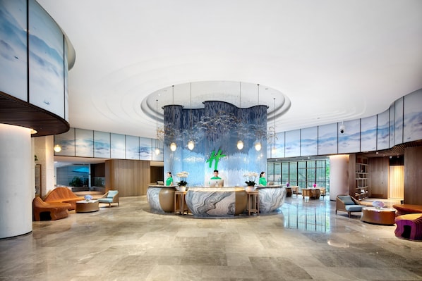 En rymlig entréhall med en central receptionsdisk, sittgrupper och ett stort fönster som ger utsikt över omgivningen.