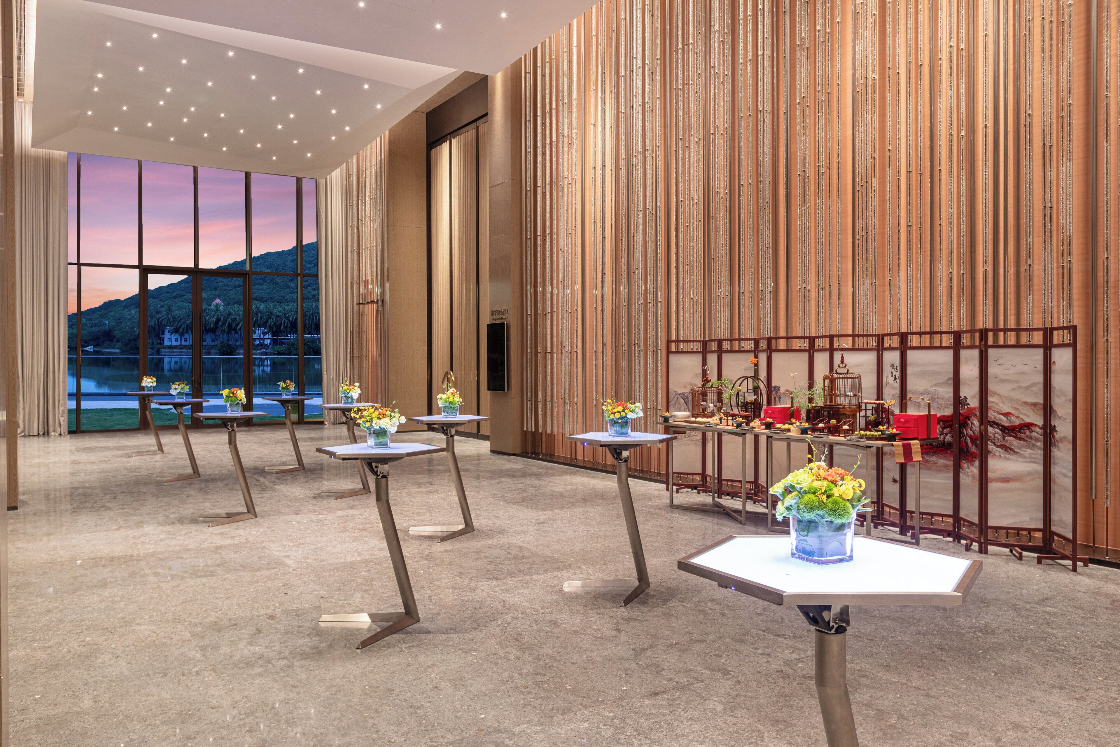 A modern hotel lobby with a large window, multiple tables with floral centerpieces, and a decorative wall with vertical lines.