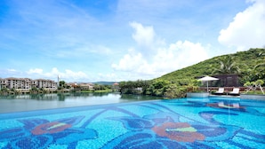 Une piscine ornée de mosaïques, entourée d’une végétation luxuriante et sous un ciel dégagé.