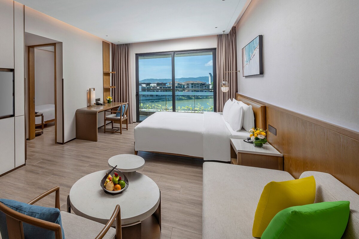 Une chambre d’hôtel avec un lit, un bureau, une télévision, une table à manger avec des fruits et une vue sur la ville.