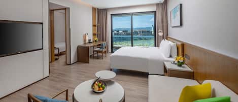 A hotel room with a bed, a desk, a TV, a dining table with fruit, and a view of the city.