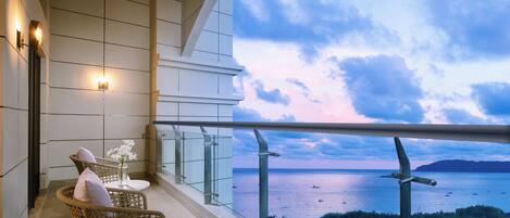 A balcony with wicker furniture, a glass railing, and a view of the sea and distant buildings.