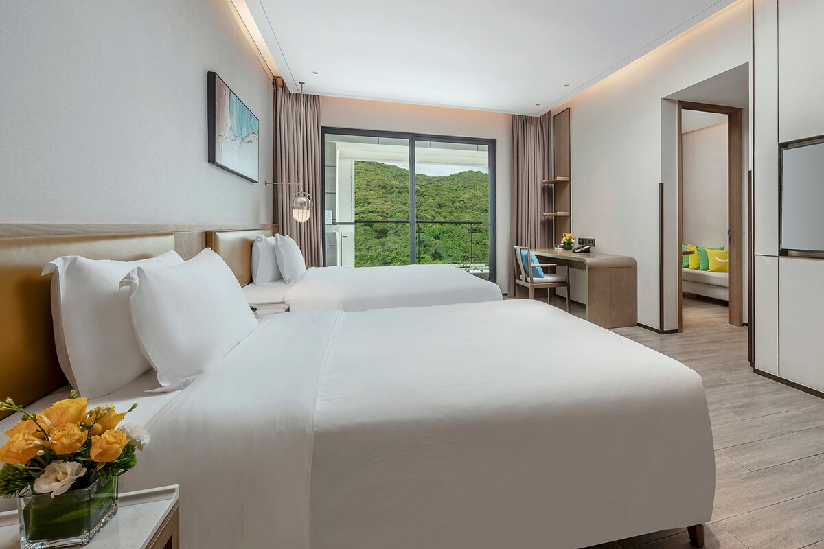 Une chambre d’hôtel avec deux lits, un bureau, une télévision et un balcon donnant sur un espace verdoyant.