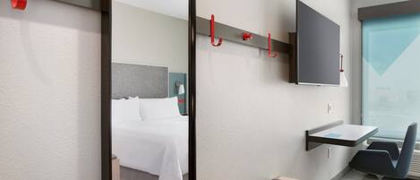 Quarto de hotel com cama, escrivaninha, cadeira, televisão fixada na parede e janela com vista.