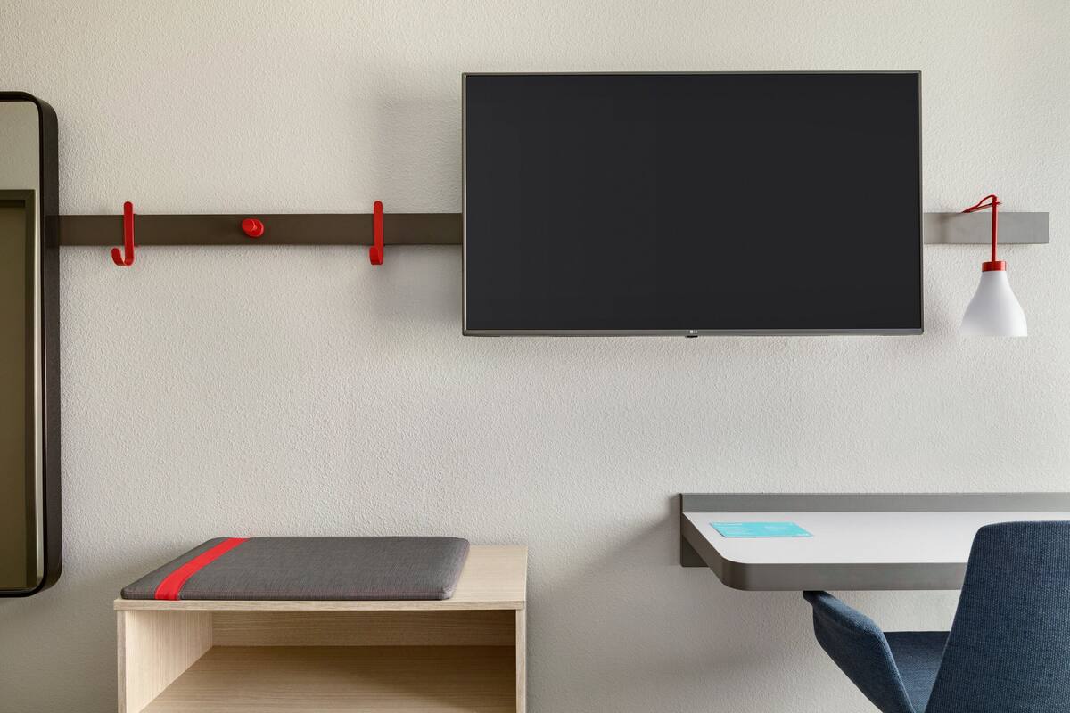 Quarto moderno com televisão de tela plana na parede, uma escrivaninha com cadeira e uma pequena estante com um almofada vermelha.