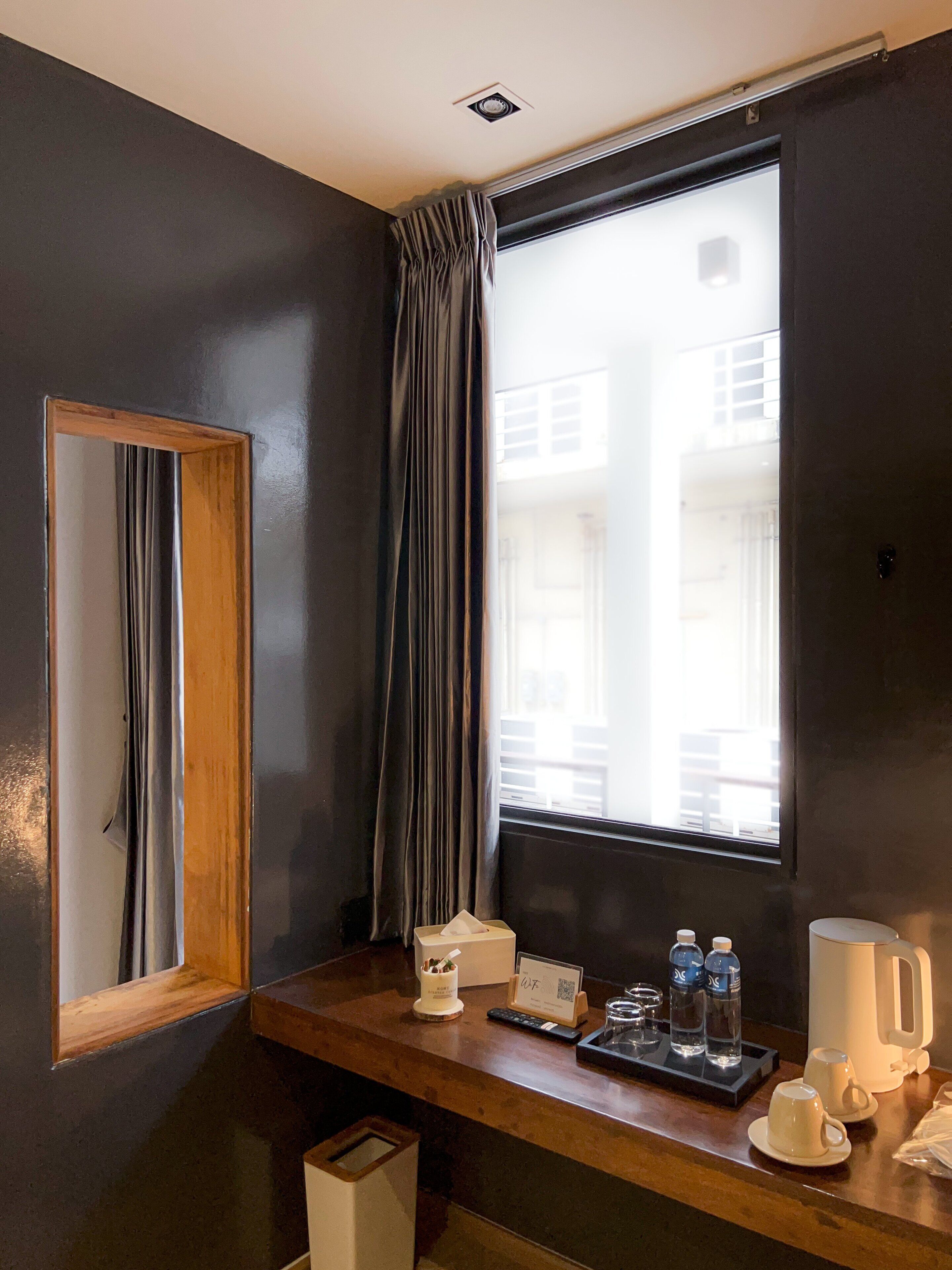 A hotel room with a wooden desk, a mirror, a window with curtains, a small table with a tissue box, a remote control, a QR code, and a coffee maker.