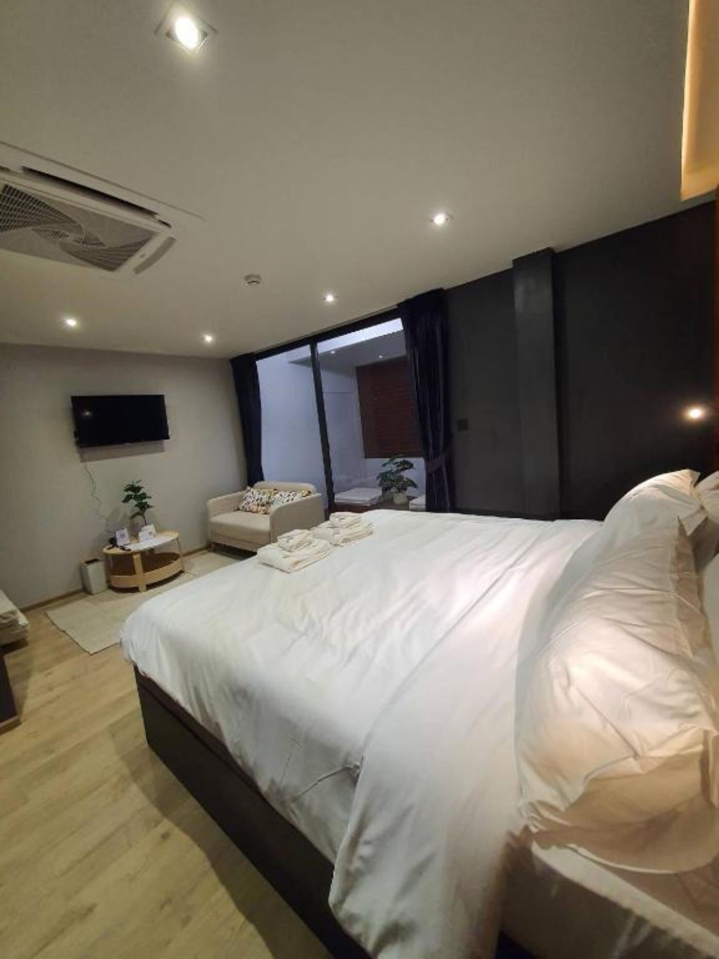 A modern hotel room with a large bed, a flat-screen TV mounted on the wall, a small seating area, and a window providing a view of the outside.