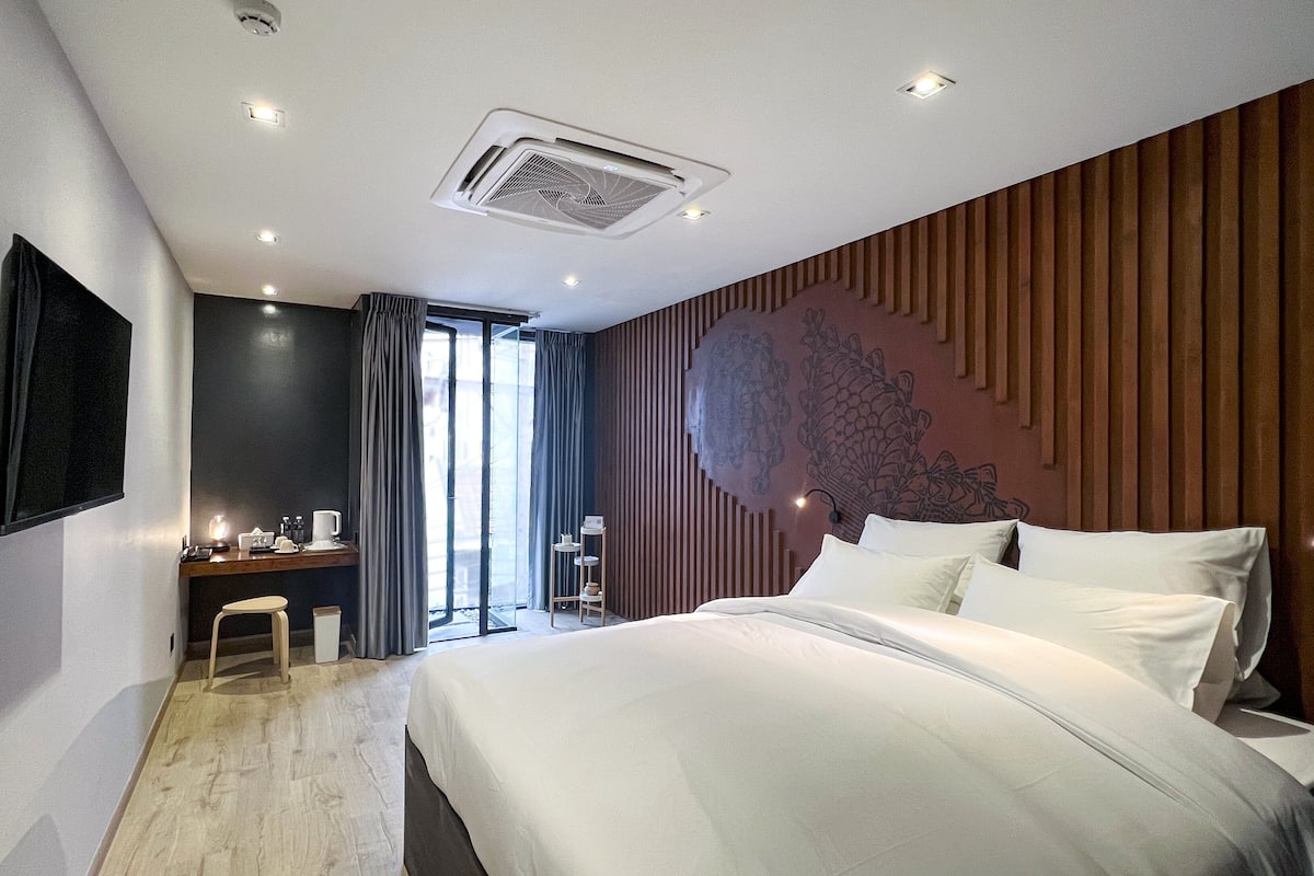 A modern hotel room with a large bed, a desk with a chair, a flat-screen TV mounted on the wall, and a large window with curtains.
