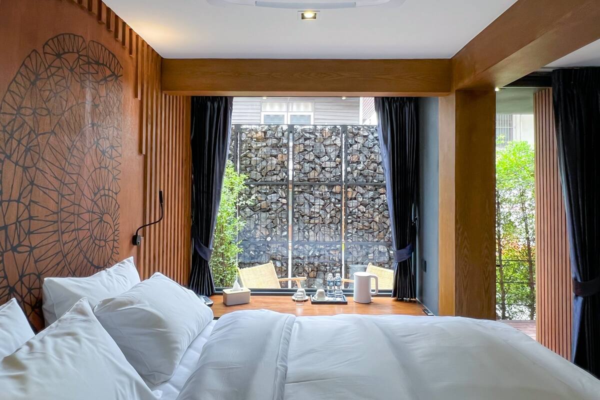 A bedroom with a large bed, a stone wall, and a view of greenery outside.