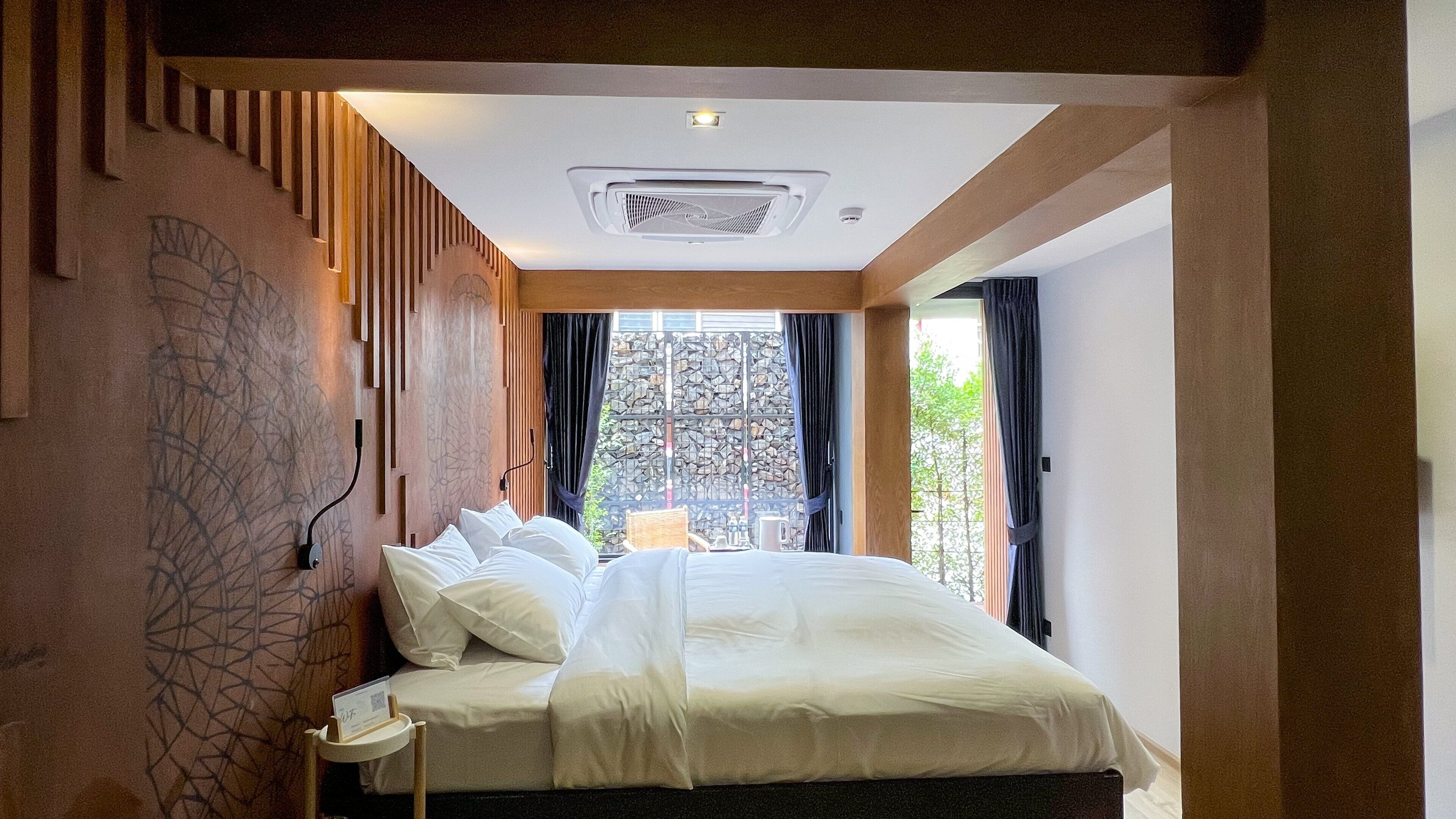 A bedroom with a bed, a window with curtains, a stone wall, and a ceiling light.