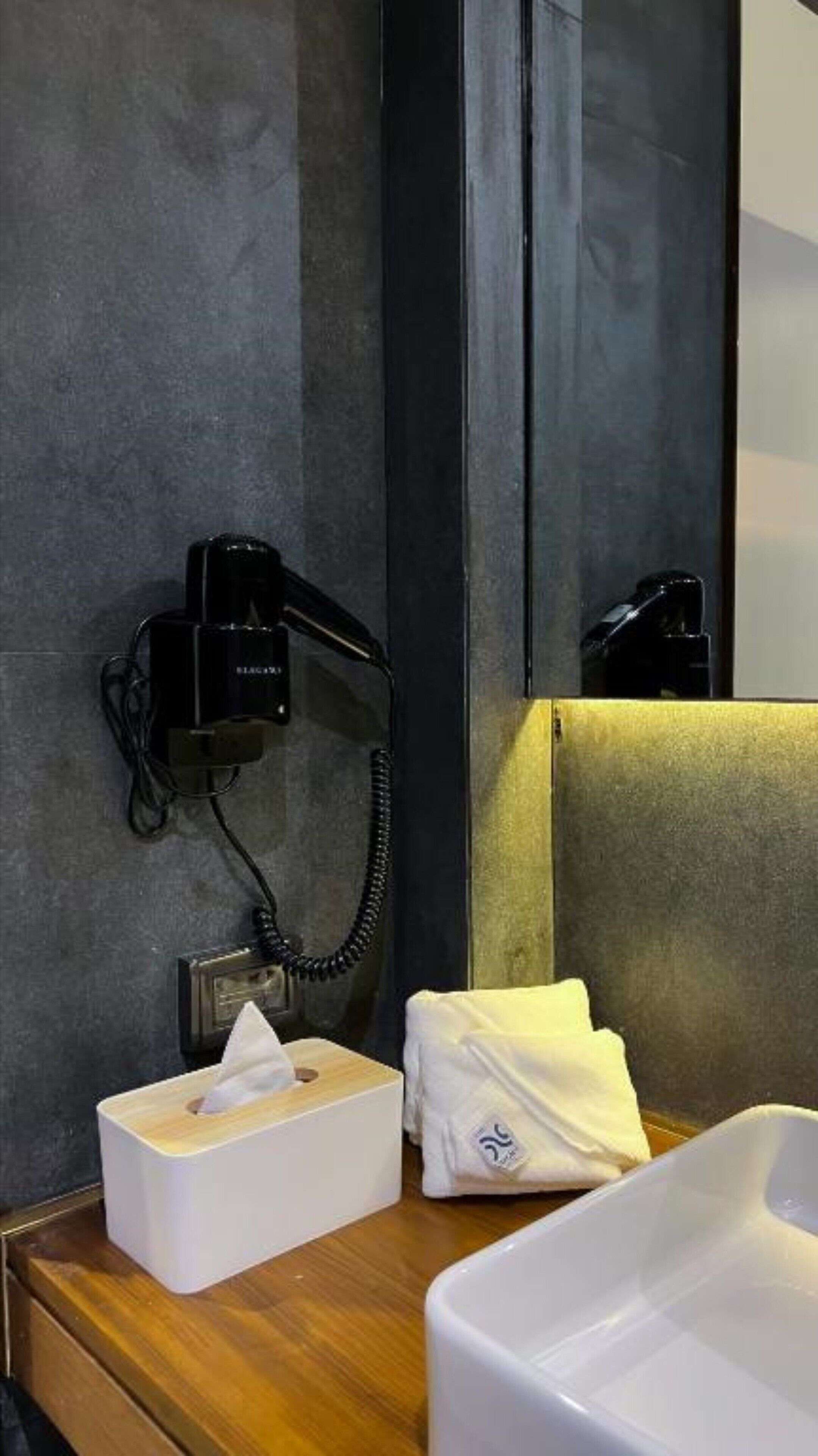 A hotel room with a black hairdryer on a wall, a tissue box, and a white towel on a wooden surface.