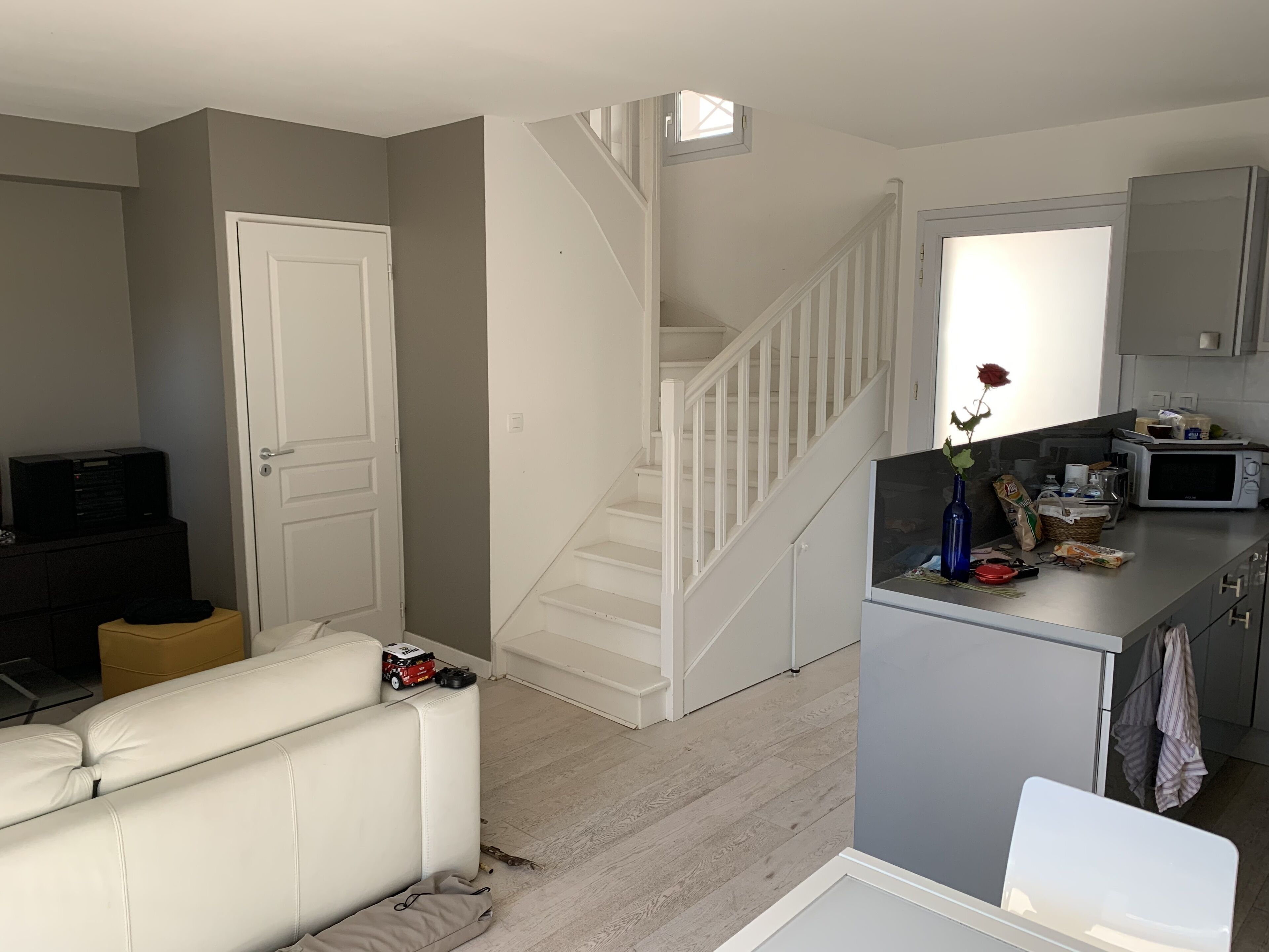 Ein Wohnzimmer mit einem weißen Sofa, einem kleinen Tisch mit einem Spielzeugauto und einer Tür. Im Hintergrund ist eine Küchenzeile mit einer Mikrowelle und einem Spülbecken zu sehen.