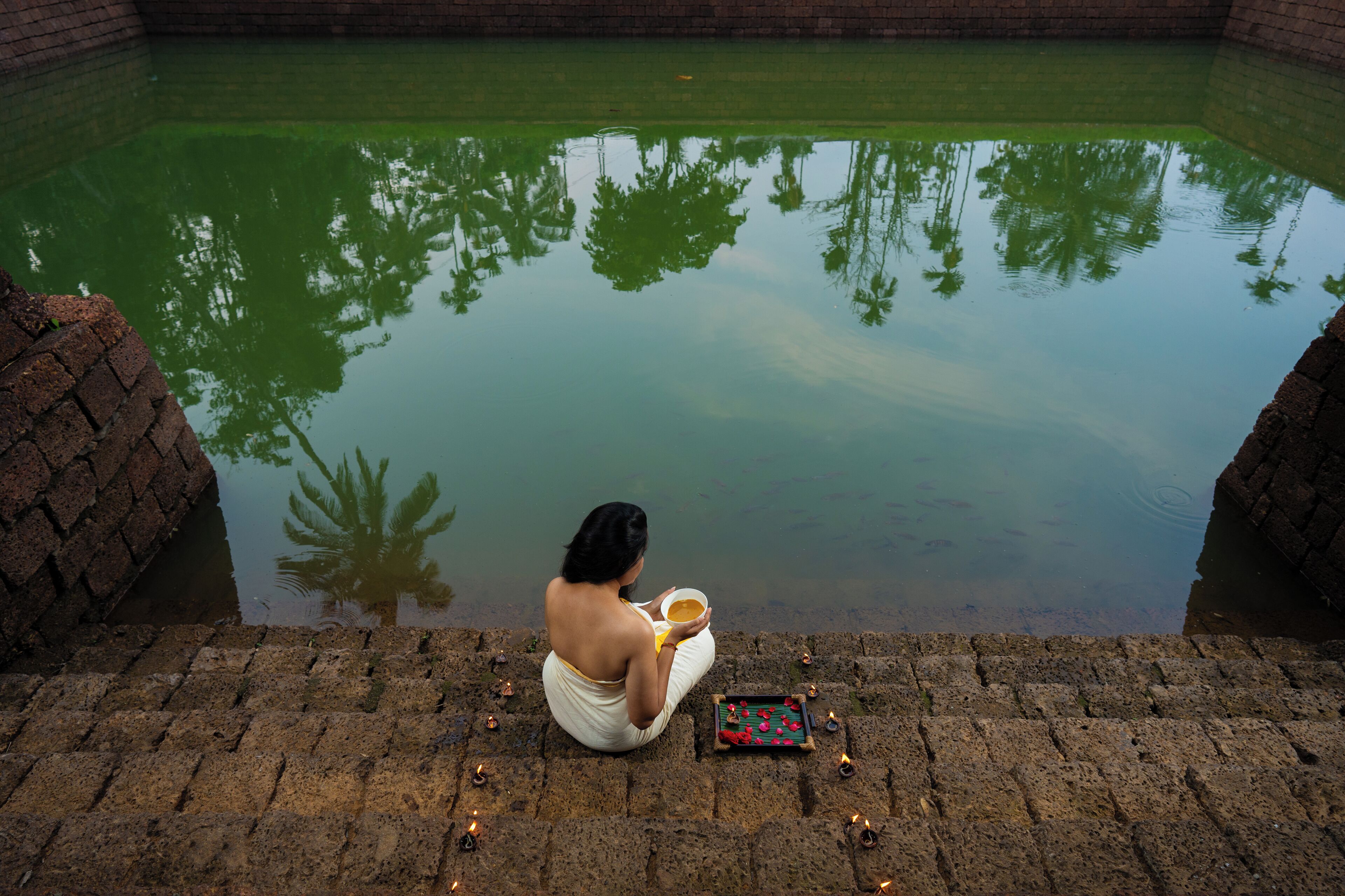 A person sitting by a reflective water body, with brick structures and a small table with items.