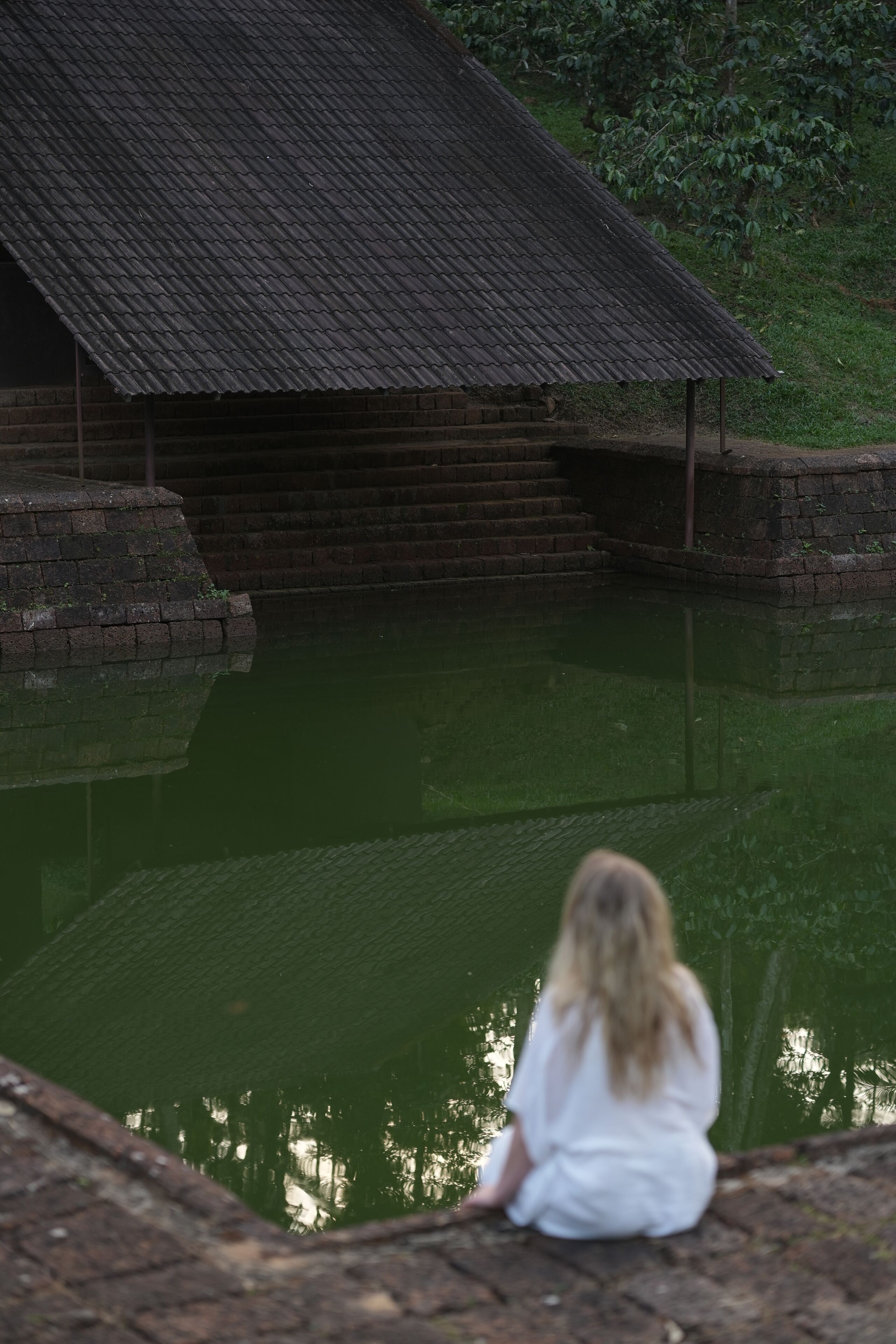 A person sitting by a calm pond with a wooden structure in the background.