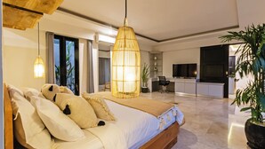 A modern bedroom with a large bed, a hanging light fixture, and a view of a kitchen and dining area.