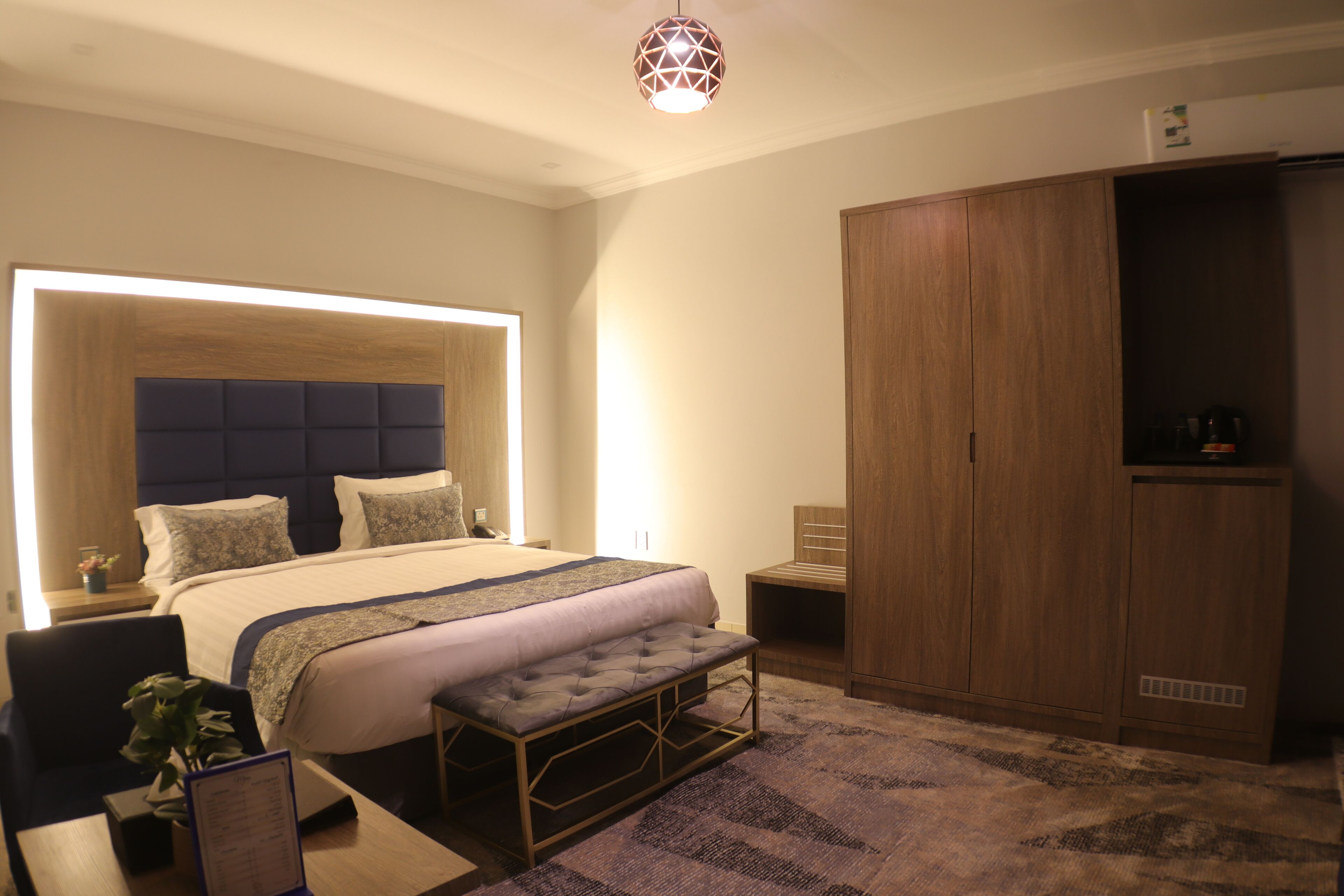 A modern bedroom with a large bed, a bedside table, a bench, a wardrobe, and a unique ceiling light fixture.