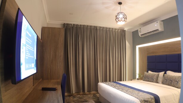 A hotel room with a large bed, a flat-screen TV mounted on the wall, a wooden desk, and a ceiling light fixture.