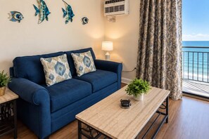 A room with a blue sofa, a wooden table, a potted plant, and a view of the ocean through a window with patterned curtains.