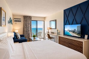 A hotel room with a large bed, a desk with a computer, a TV, and a balcony with a view of the ocean.
