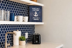Une cuisine avec un revêtement mural bleu en forme d’hexagones, une machine à café, une plante en pot et une étagère ornée d’objets décoratifs et d’une affiche motivante.