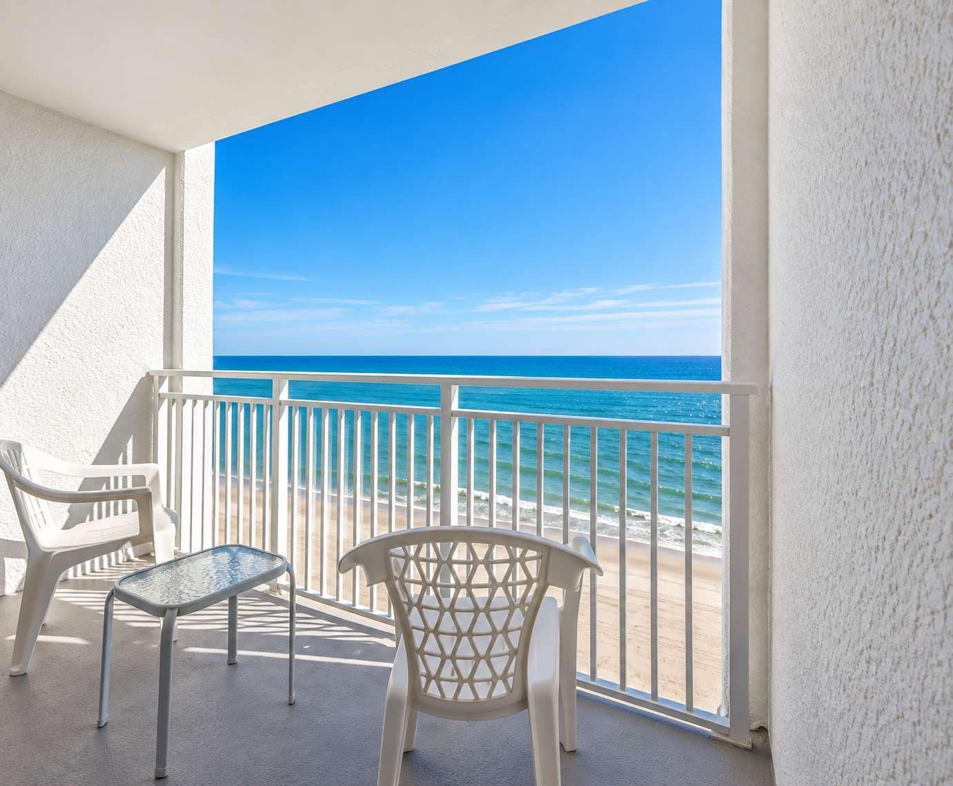A balcony with a view of the ocean, two white plastic chairs, and a glass table.