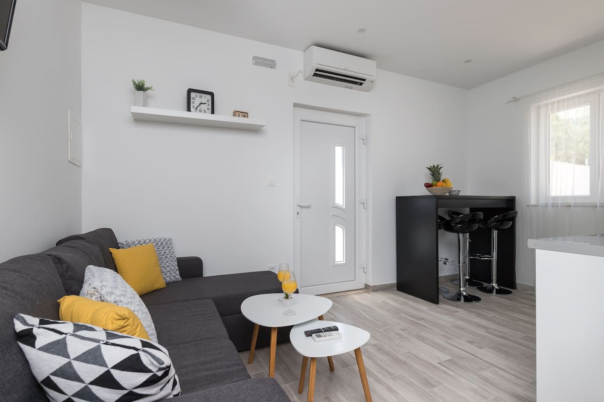 A modern living room with a grey sofa, a small white table, and a kitchen area with a bar stool.