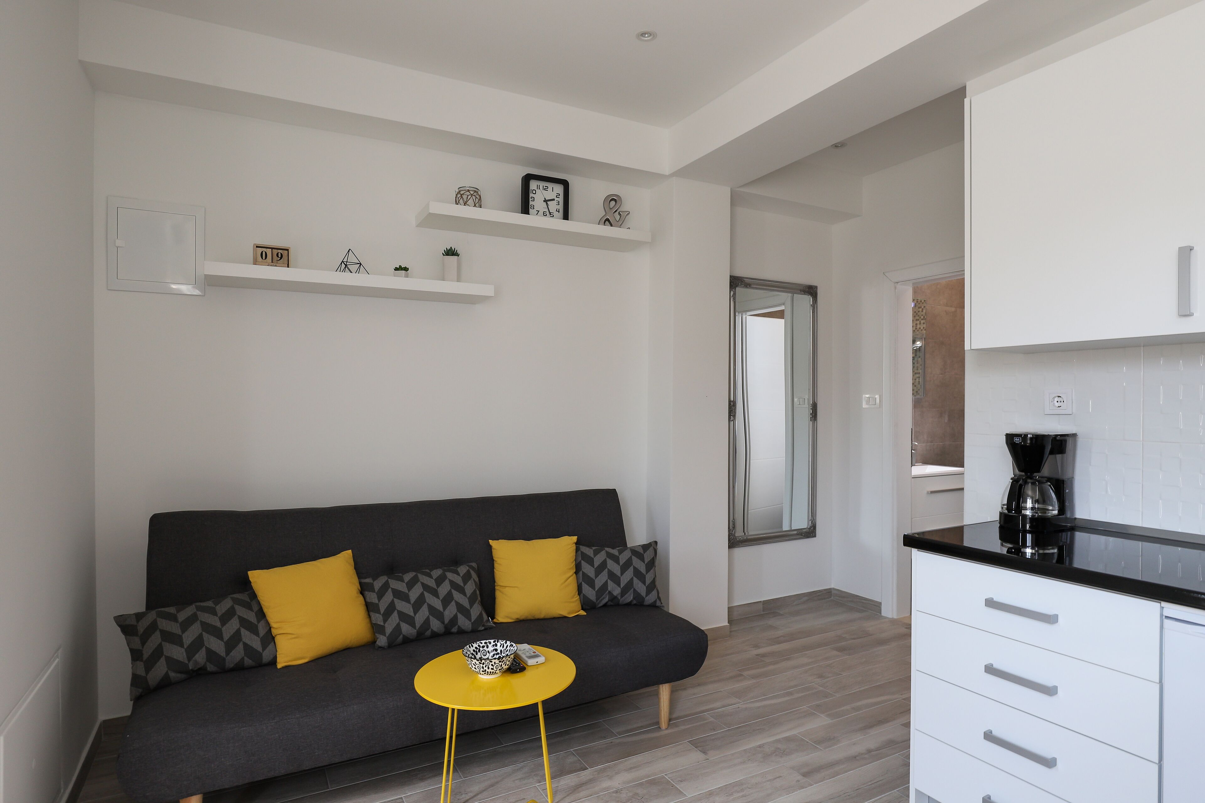 A modern living room with a grey sofa, yellow cushions, a small yellow table, and a kitchen area with white cabinets and a black countertop.