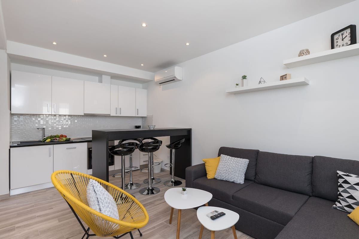 A modern living space with a grey sofa, yellow chair, and a small white table. There is a kitchen area with white cabinets and a black countertop.