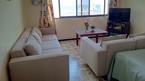 A living room with a sofa set, a coffee table with a vase, a television on a stand, and a window with a view of the sea. - Vers Les Nuages, Old Souk, Sea View Flat & Balcony (Batroun)