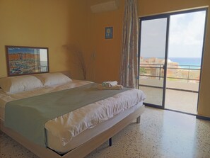 Une chambre d’hôtel avec un lit, un tableau, une plante en pot et une vue sur la mer depuis un balcon.