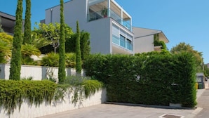 Um edifício moderno de dois andares com varanda, rodeado por vegetação exuberante e árvores altas.