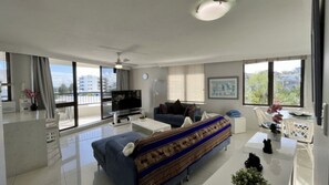 A modern living room with a sofa, a coffee table, a TV, and a balcony with a view of buildings. - Oogarding - Panoramic Views, 2BR Apt, Walk to Beach (Caloundra)