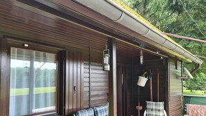 A wooden cabin with a porch, a table, two chairs, a hanging planter, and a window with curtains. - Idyllic vacation home in the middle of nature (Neunkirchen)