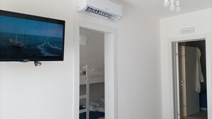 Une chambre avec un lit superposé, un appareil de climatisation, un téléviseur diffusant une image d’un bateau sur la mer, et un couloir menant à une autre chambre.