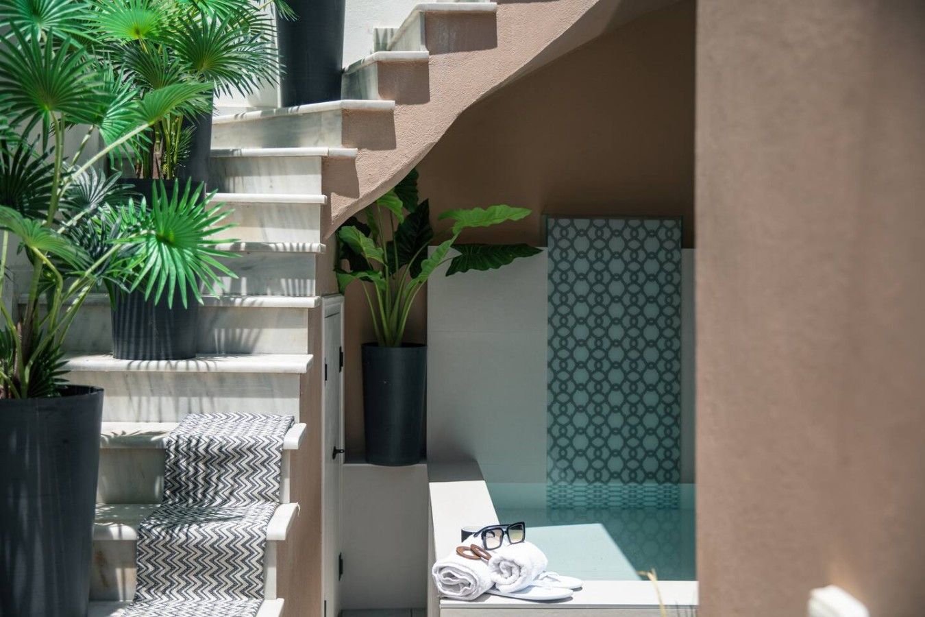 Un escalier agrémenté de plantes en pot, un mur orné de motifs et un aperçu d’une pièce où l’on aperçoit un lit et des serviettes.