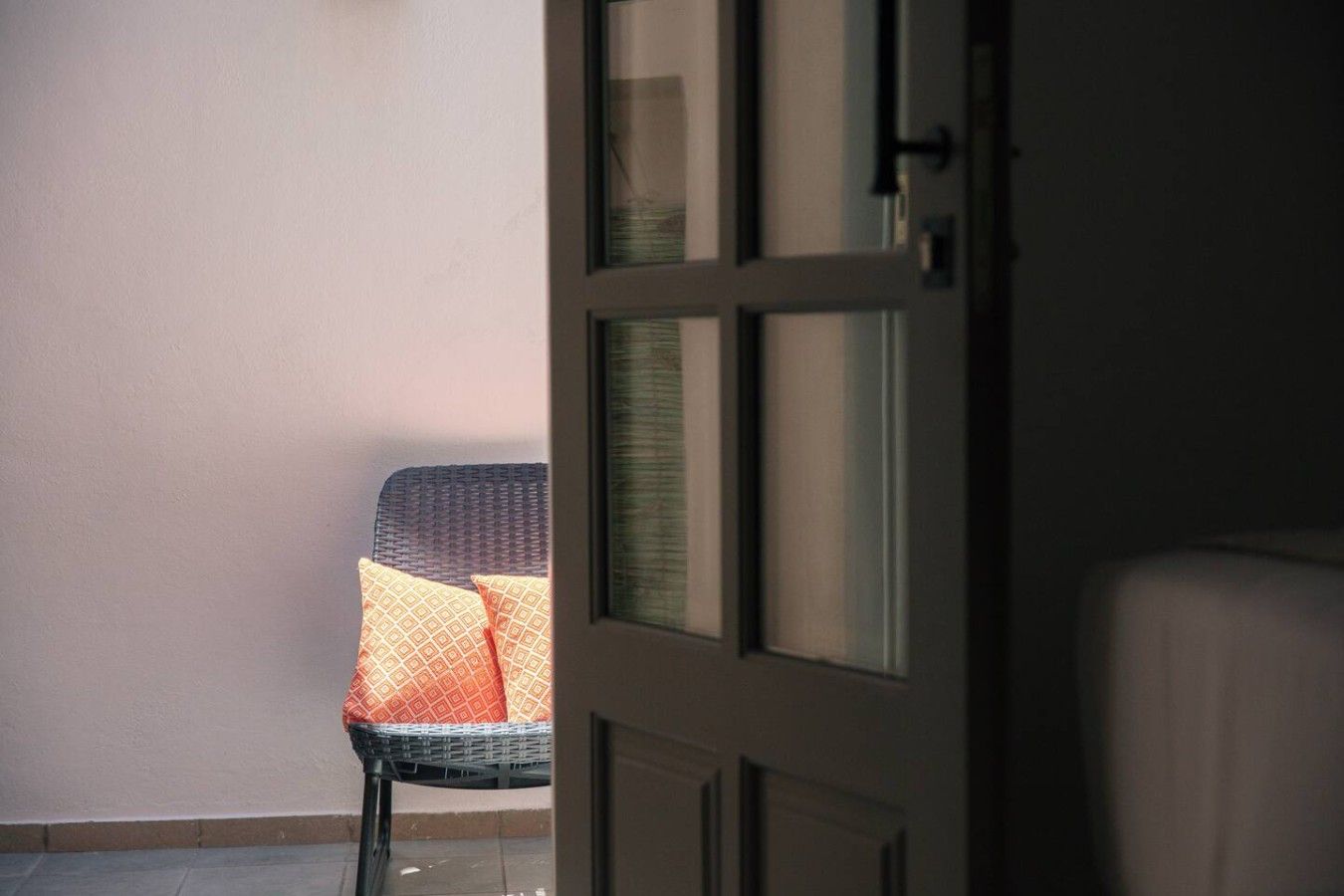 Ein Zimmer mit einem Korbstuhl in der Nähe eines Fensters, einer geschlossenen Tür und einer schlichten Wand.