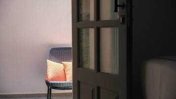 Ein Zimmer mit einem Korbstuhl in der Nähe eines Fensters, einer geschlossenen Tür und einer schlichten Wand.