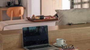 Una habitación moderna con un escritorio de madera, una computadora portátil, una taza y un par de lentes. Se ve una cama con un almohadón y un armario de madera al fondo.