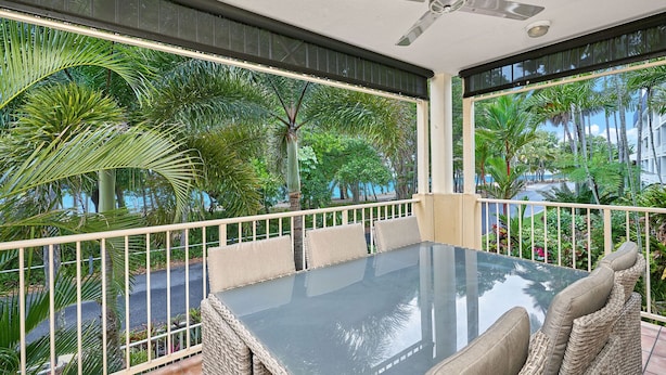 Un balcon avec une table en verre et des chaises en osier, entouré d’une végétation tropicale luxuriante et offrant une vue sur un immeuble.