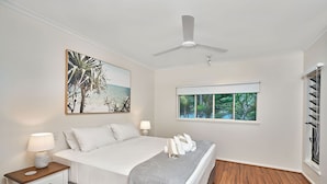 Une chambre à coucher comprenant un lit, des tables de chevet, un ventilateur de plafond, une fenêtre donnant sur un paysage verdoyant et un tableau accroché au mur.
