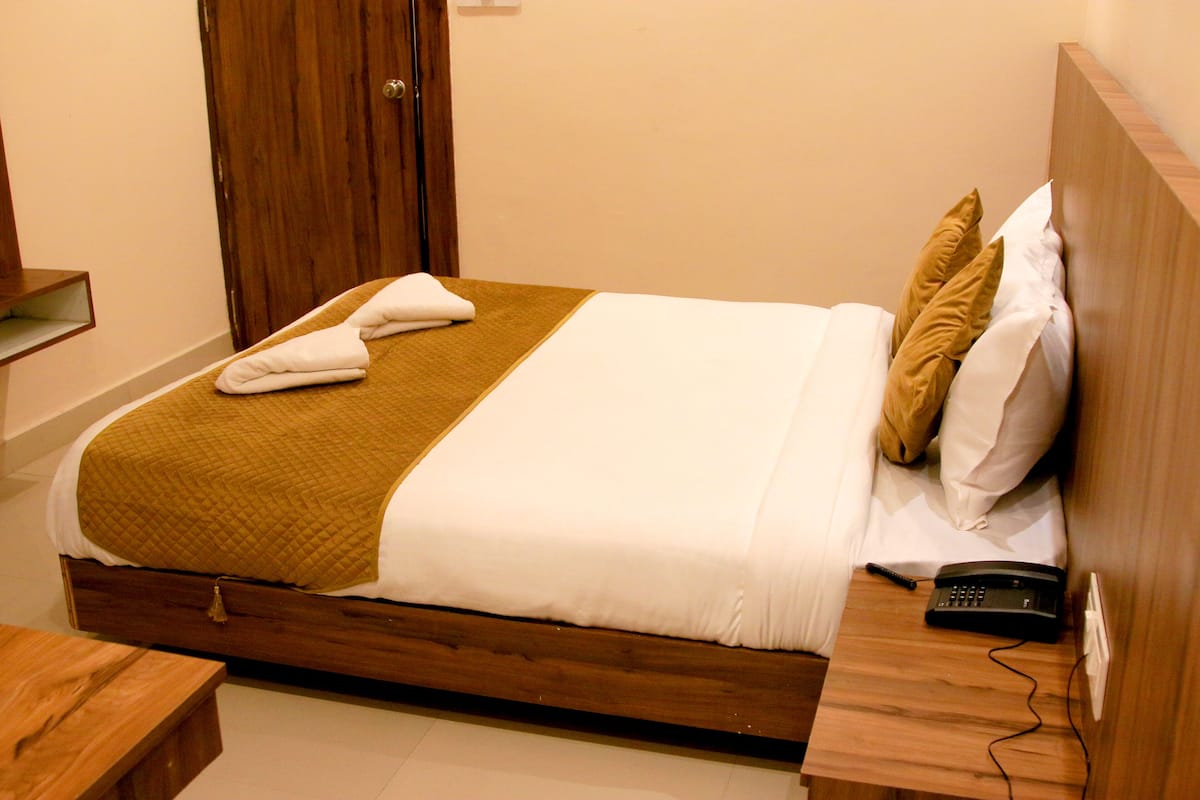 A neatly made bed with a brown quilt, white linens, and two towels folded at the foot. A remote control is placed on the wooden bedside table.