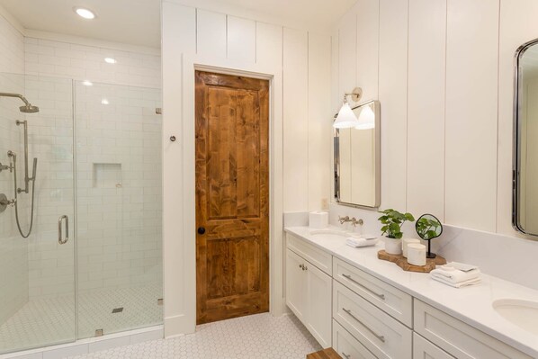 Une salle de bain moderne avec une douche à parois de verre, une porte en bois, des meubles blancs, un grand miroir et une petite plante posée sur le meuble-lavabo.